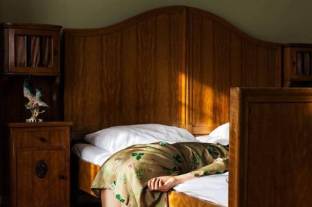 dans la chambre ensoleillée, déjà habillée, une femme agenouillée sur le sol repose encore un peu le haut de son corps en appuyant sa tête sur le lit
