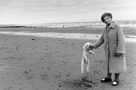 sur une plage déserte, une vieille femme en manteau d'hiver, une laisse à la main,  pose en train de faire sauter son caniche