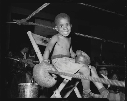 assis dans le coin d'un ring, un très jeune garçon noir portant d'énormes gants de boxe