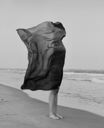 une femme sur la plage, le voile de ses vêtements plaqué sur l'ensemble de son corps -tête comprise - par un coup de vent marin