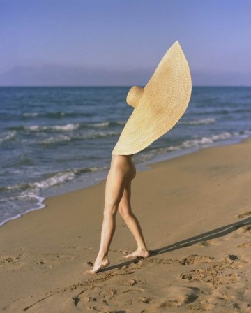 sur la plage, une femme nue face à la mer, une immense capeline plaquée par le vent sur son visage