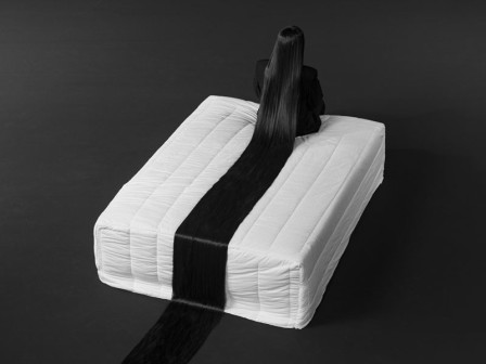 étalée dans le sens de sa longueur, la très longue chevelure brune d'une femme assise sur un lit trace une ligne de partage sur un matelas blanc