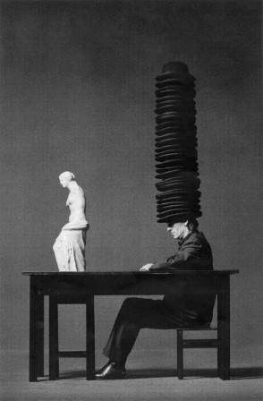 assis à une table sur laquelle est posée une statue, un homme porte sur sa tête toute une pile de chapeaux