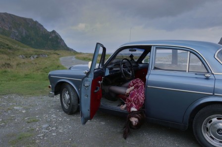 sur un chemin de bord de mer, un ancien modèle de Volvo bleue arrêtée, le corps de la conductrice brune pendant par la porte ouverte, la tête près du gravier, les yeux ouverts